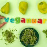 A green table featuring vintage magnetic fridge letters in various colours that spell out VEGANUARY. There is a selection of green vegetables and herbs surrounding the lettering